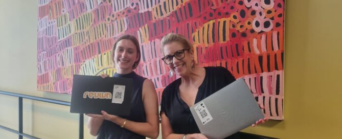 Two women smiling, holding laptops with stickers, stand in front of a vibrant abstract painting. The atmosphere is cheerful and lively.