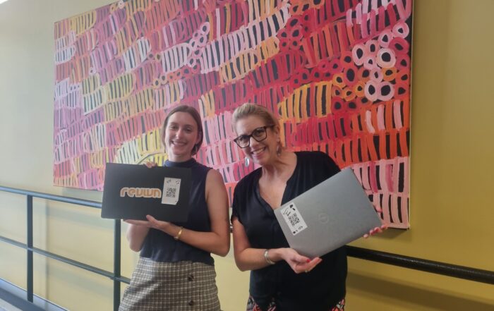 Two women smiling, holding laptops with stickers, stand in front of a vibrant abstract painting. The atmosphere is cheerful and lively.