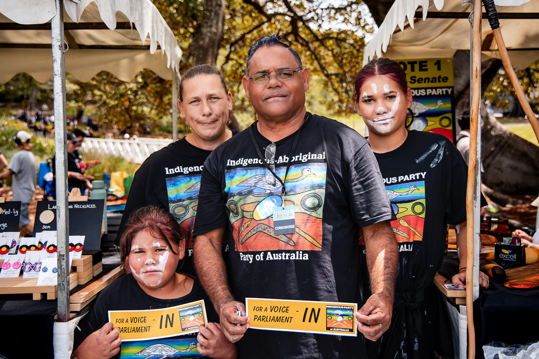 A group of four people, wearing "Indigenous Party of Australia" shirts, stand under a tent, smiling and holding "For a Voice in Parliament" signs.