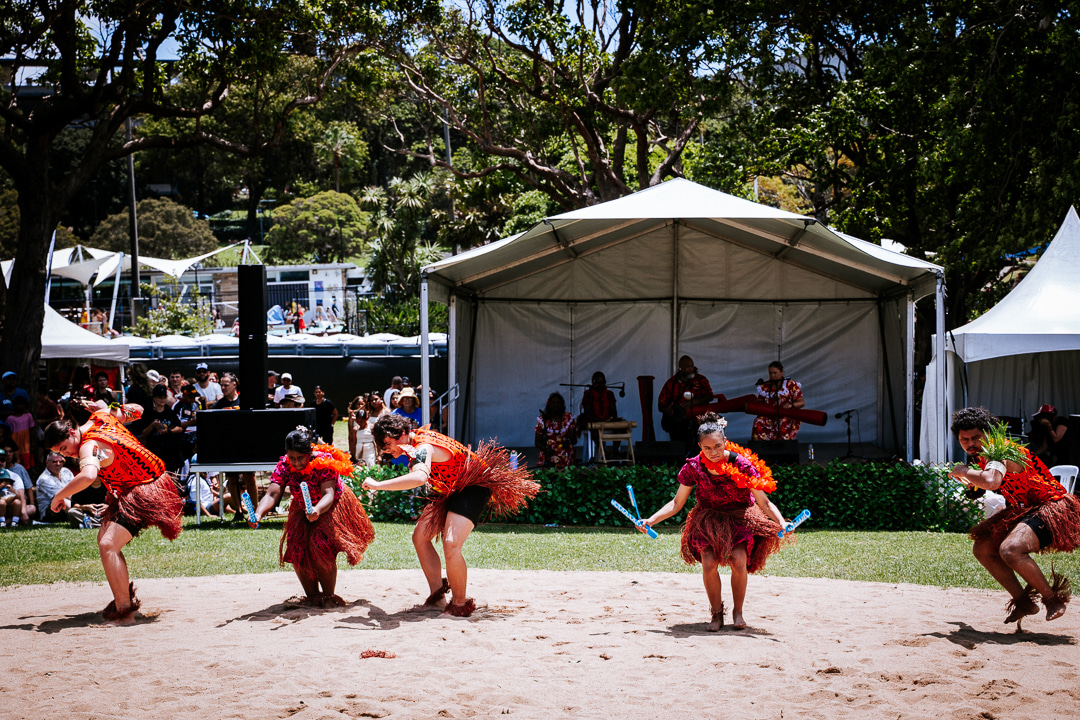 A group of dancers in vibrant red and orange traditional attire perform energetically on a sandy stage, backed by musicians under a tent, with a lively audience and lush trees surrounding them.