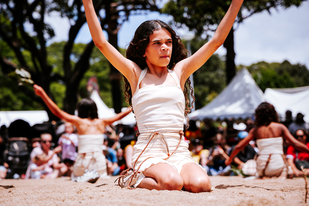 A dancer in a light-colored dress performs on the sand, surrounded by an audience at an outdoor festival, with trees in the background.