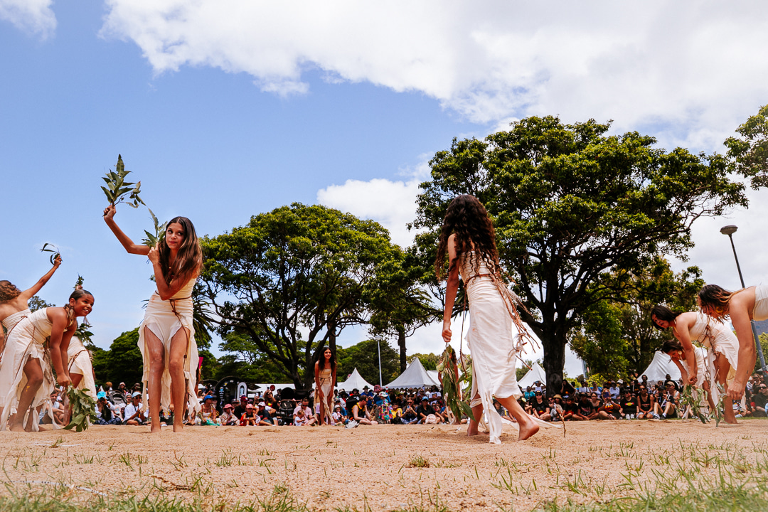 Dancers in traditional attire perform with leaves in a sunny park, surrounded by a large audience and trees. The scene is vibrant and cultural.