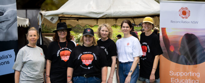 Two images of Indigenous Australian individuals holding a "Vote Yes" poster. They are outdoors with clear skies and dry landscape, conveying a hopeful message.