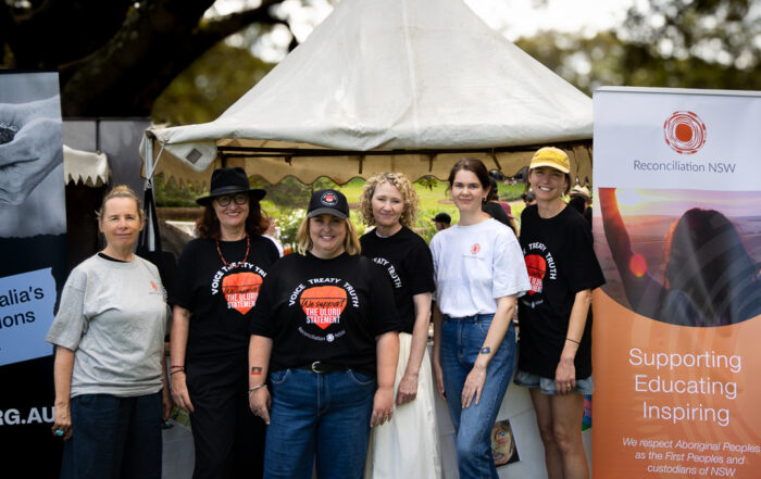 Two images of Indigenous Australian individuals holding a "Vote Yes" poster. They are outdoors with clear skies and dry landscape, conveying a hopeful message.