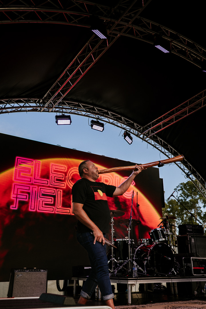 A performer holds a Yidaki on stage, with vibrant "Electric Fields" lighting and musical instruments in the background.