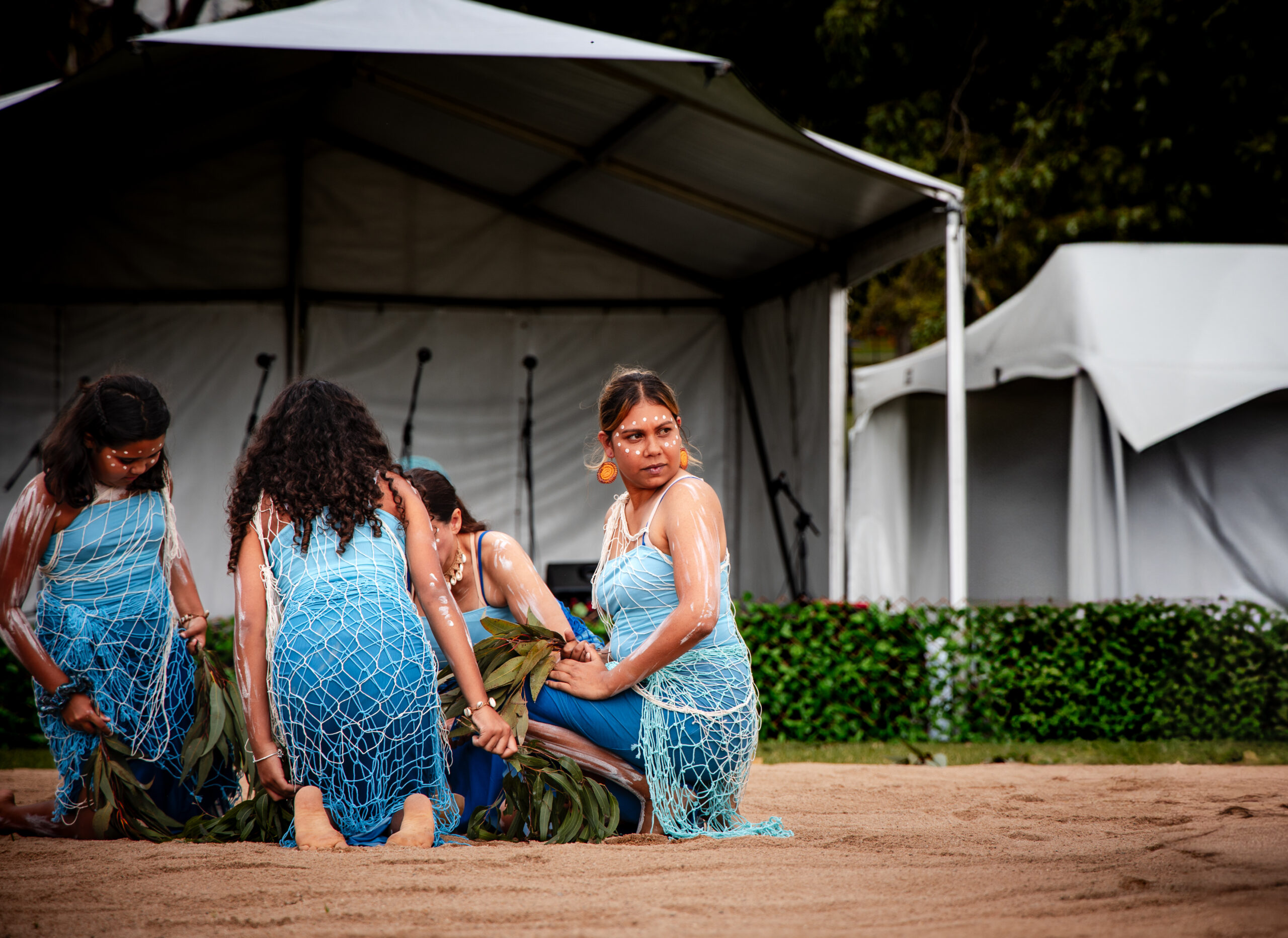 Four women kneel on sandy ground, performing a cultural dance. They wear blue attire with netted overlays and body paint. A canopy and foliage are in the background.