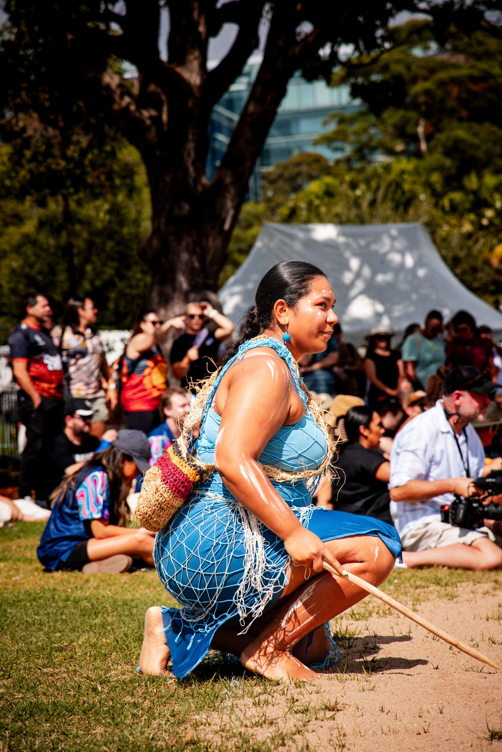 A woman in a blue traditional outfit kneels on grass, holding a stick, with a crowd and tent in the background. The scene is vibrant and lively.