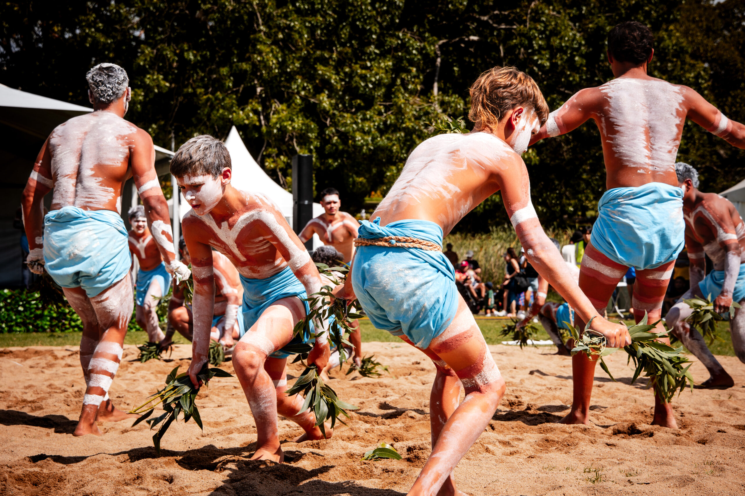 A group of people, painted with white patterns, dance energetically on sandy ground, holding green branches. They wear blue cloths, conveying a joyous cultural celebration outdoors.