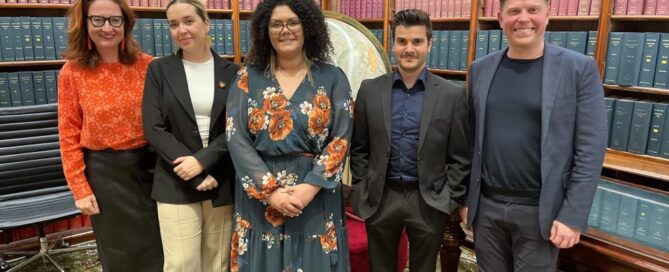 A group of five people stand smiling in a library with shelves of books. They wear business attire, and the setting is formal yet warm and welcoming.