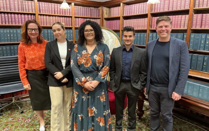 A group of five people stand smiling in a library with shelves of books. They wear business attire, and the setting is formal yet warm and welcoming.