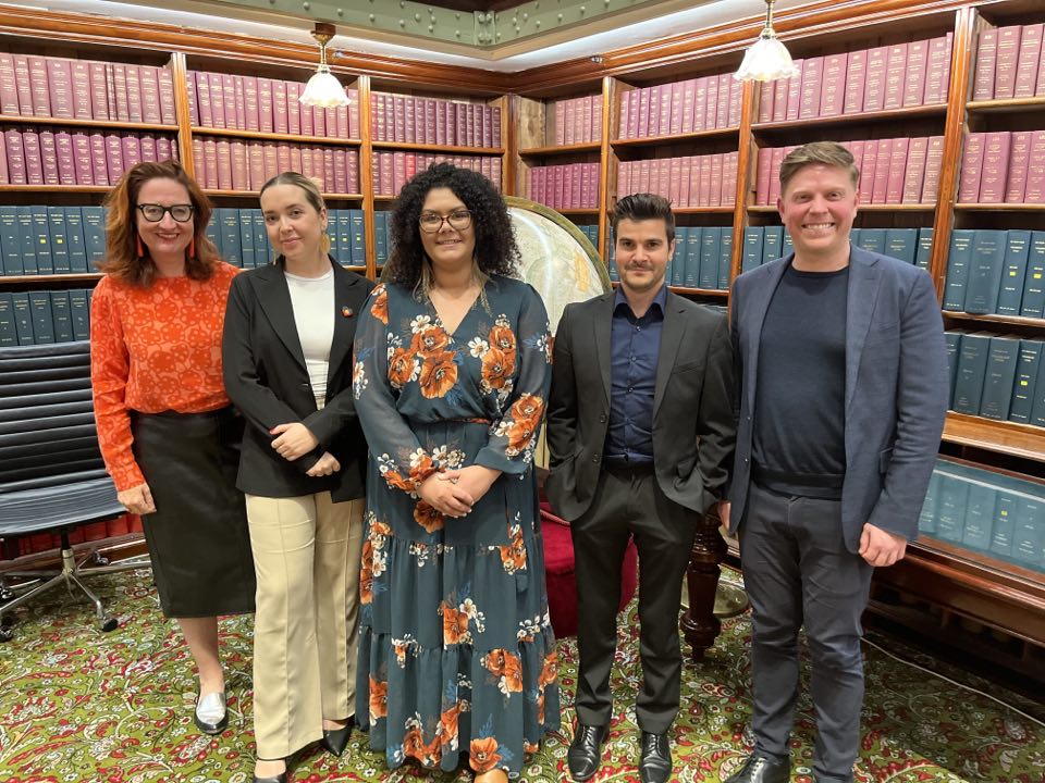 A group of five people stand smiling in a library with shelves of books. They wear business attire, and the setting is formal yet warm and welcoming.