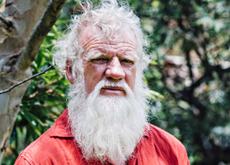 Elderly man with a long white beard and curly hair stands outside, wearing a red shirt. The background is lush with green foliage, creating a natural setting.