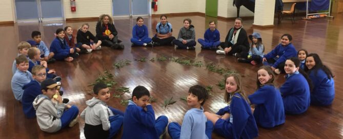 A diverse group of children and two adults sit in a circle on a polished wooden floor, surrounding eucalyptus leaves. They appear engaged and happy.