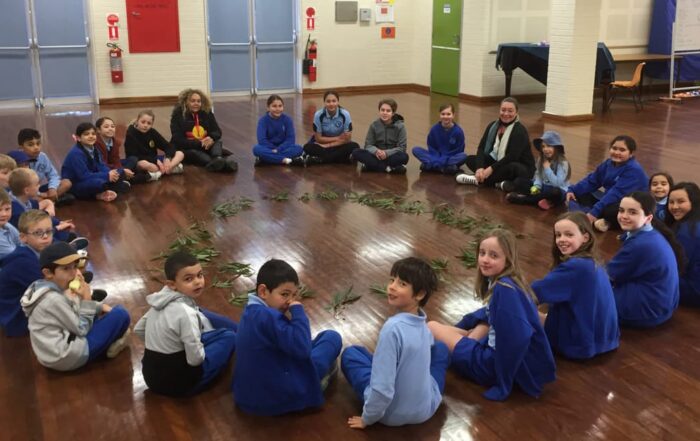 A diverse group of children and two adults sit in a circle on a polished wooden floor, surrounding eucalyptus leaves. They appear engaged and happy.