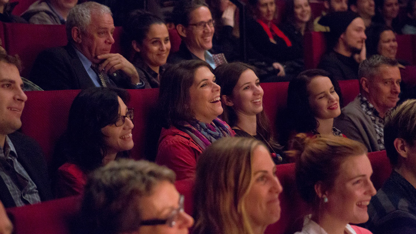 Audience members seated in a theater with red chairs, smiling and laughing, creating a joyful and lively atmosphere during a performance.