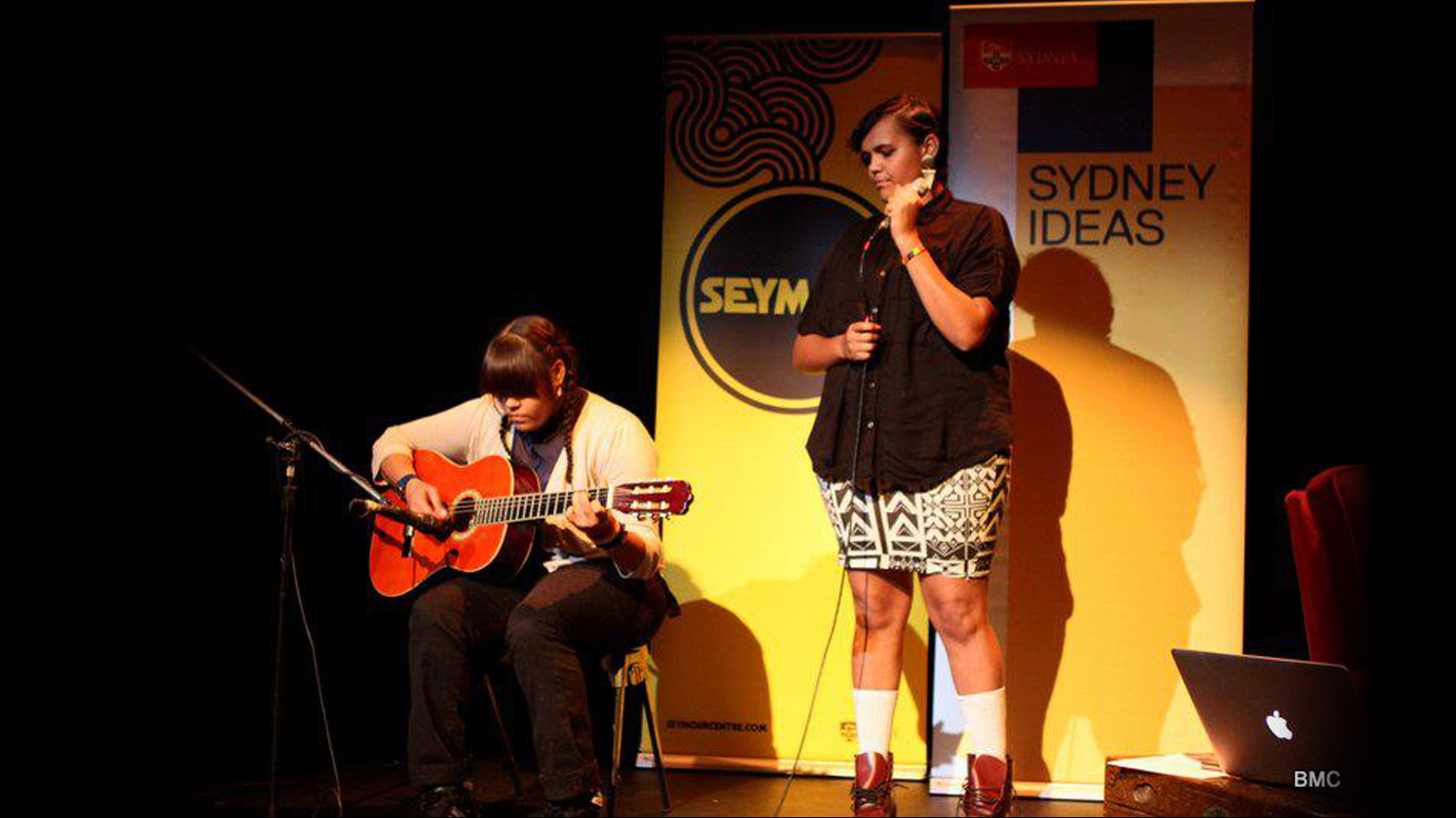 Two musicians perform on stage; a seated guitarist in a sweater and a standing singer with a microphone. "Sydney Ideas" and "SEYM" banners are in the background.