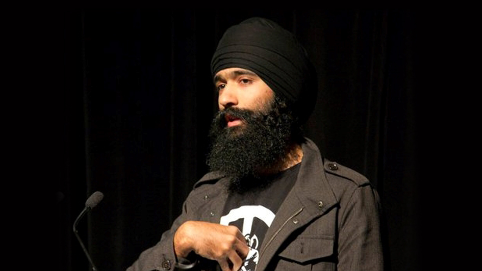 Man wearing a black turban and beard speaks passionately at a conference, standing against a dark background. He gestures with one hand on his chest.