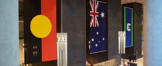 Large interior space displaying three vertical flags: Aboriginal, Australian, and Torres Strait Islander. Modern lighting, dark walls. Majestic, respectful tone.