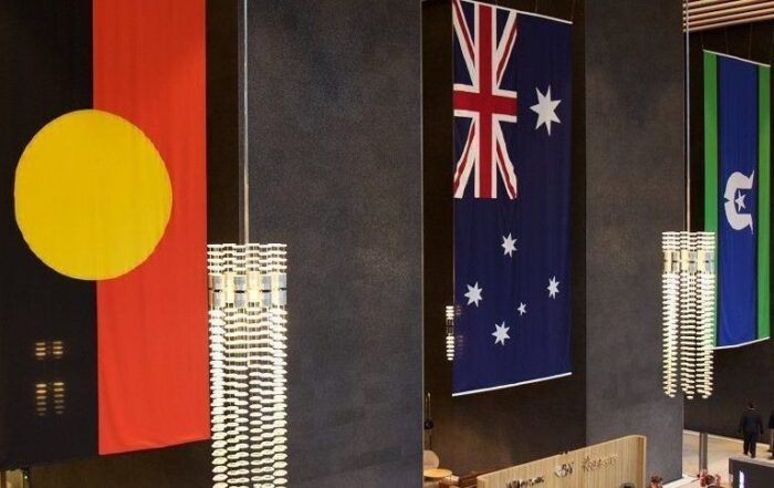 Large interior space displaying three vertical flags: Aboriginal, Australian, and Torres Strait Islander. Modern lighting, dark walls. Majestic, respectful tone.