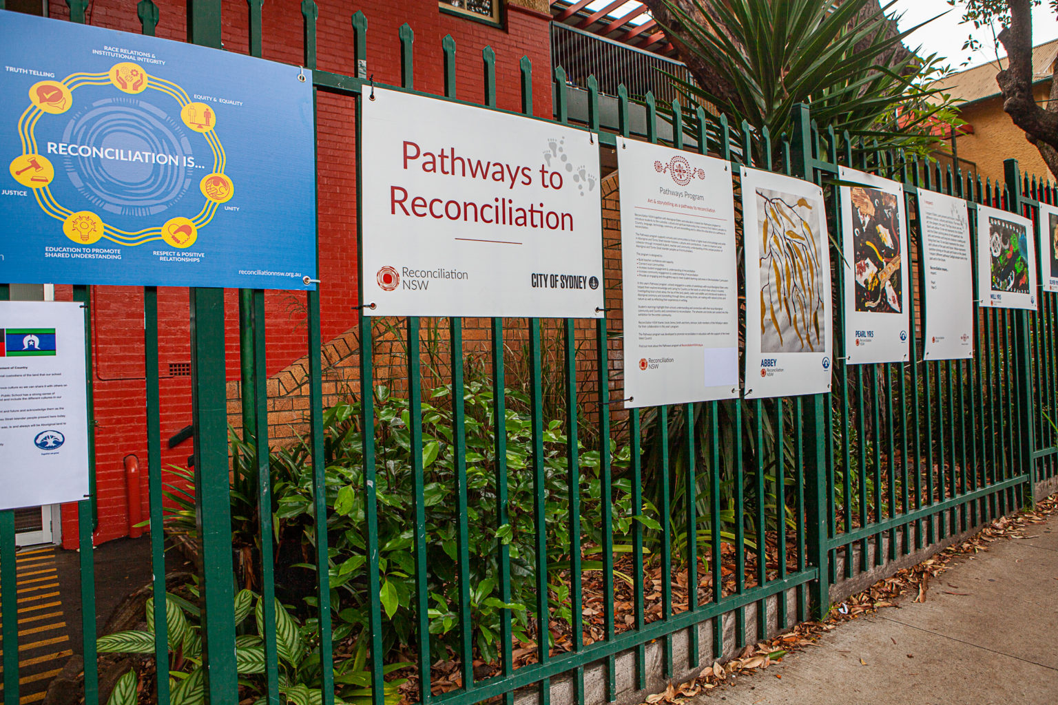 A series of educational posters about reconciliation hangs on a green fence. The setting is outdoors, near red and orange brick walls, conveying a message of unity and understanding.