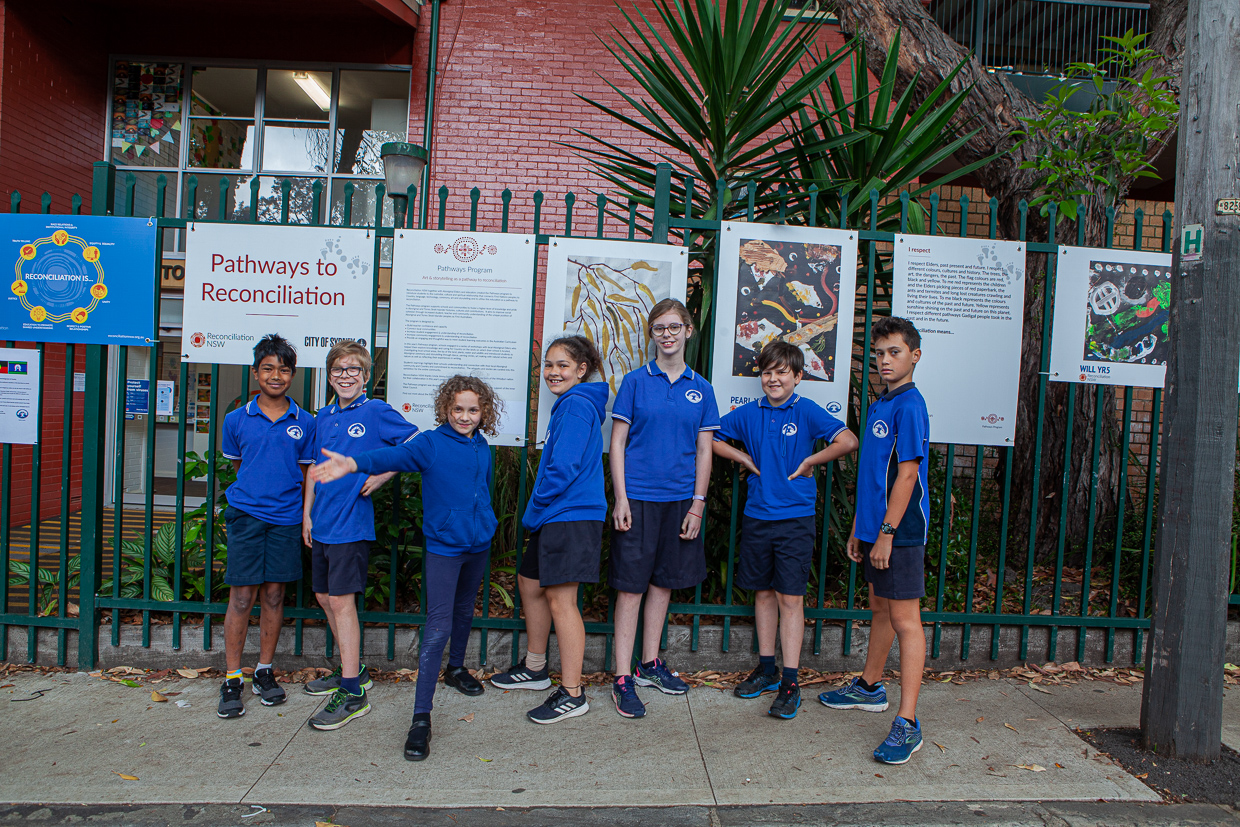 Seven children in blue shirts stand in front of a school fence displaying reconciliation-themed posters. They pose playfully, exuding a cheerful and inclusive atmosphere.