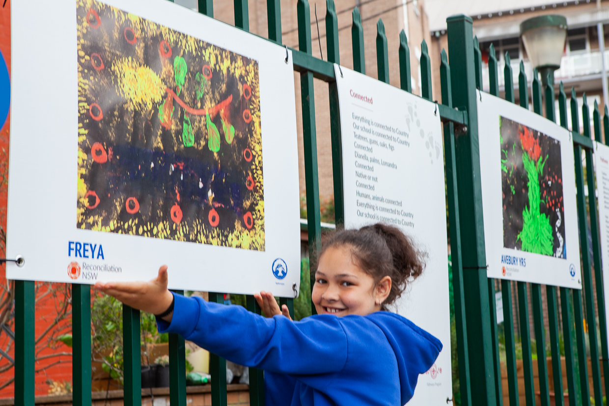 Smiling child in a blue hoodie gestures toward colorful, abstract artwork displayed on a fence. Art is vibrant with reds, greens, and yellows.