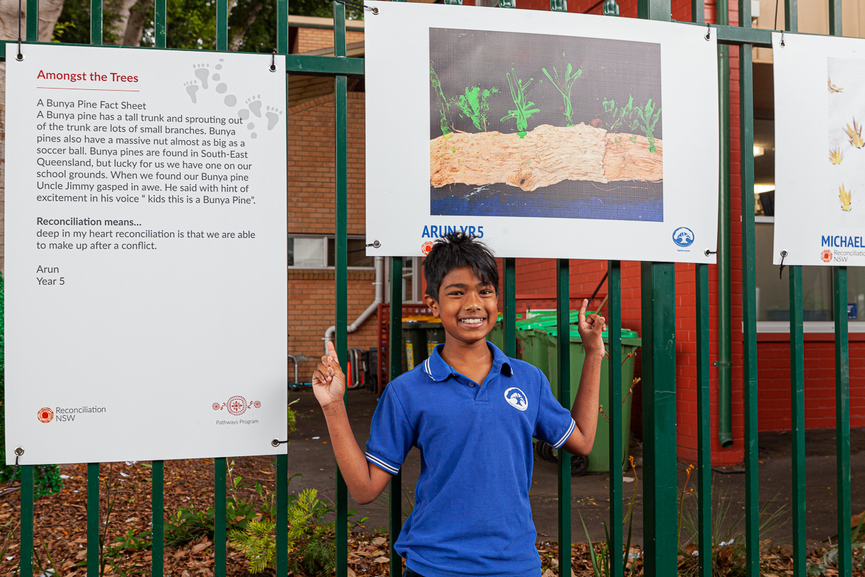 Young boy in a blue shirt smiles and points at an art display on a fence. The left poster has text about Bunya pines and reconciliation. The setting is cheerful and educational.