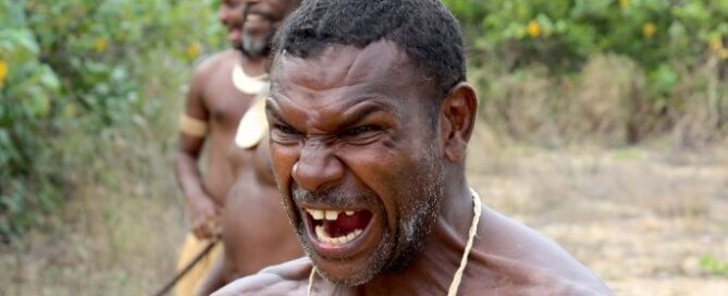 A man with a fierce expression stands in the foreground, mouth open wide. Behind him, two men are visible, surrounded by lush greenery, conveying intensity.