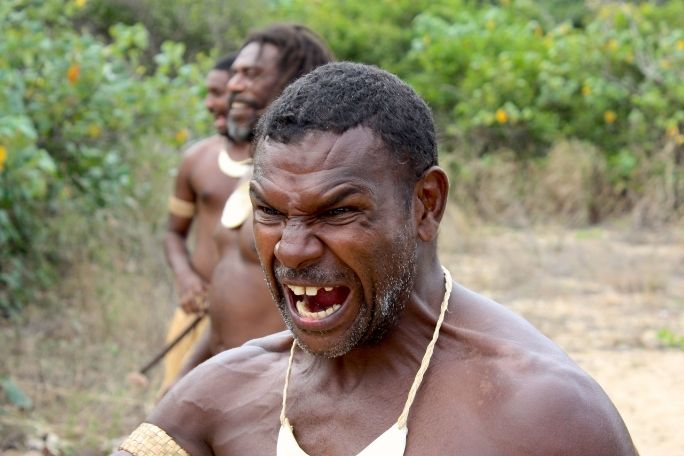 A man with a fierce expression stands in the foreground, mouth open wide. Behind him, two men are visible, surrounded by lush greenery, conveying intensity.