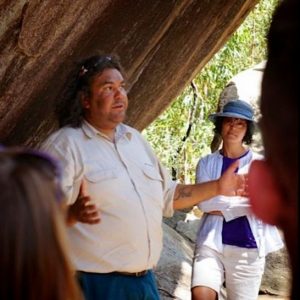 An individual speaks expressively to a small group gathered under a large rock formation. An attentive listener stands nearby, showcasing an outdoor learning setting.