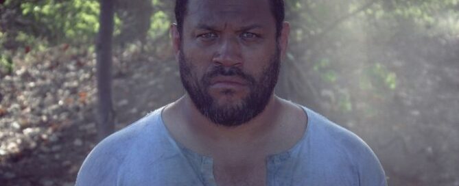 A man with short hair and a beard stands in a sunlit forest, wearing a weathered shirt. He has a serious expression, with sunbeams filtering through trees.