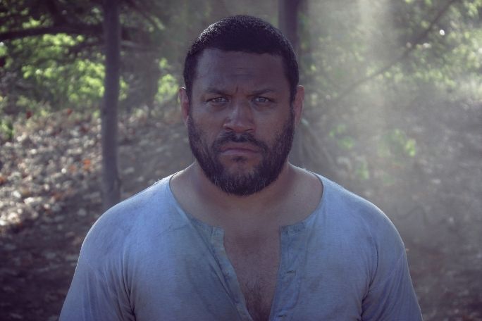 A man with short hair and a beard stands in a sunlit forest, wearing a weathered shirt. He has a serious expression, with sunbeams filtering through trees.