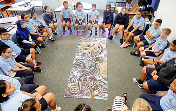 A diverse group of students in blue uniforms sit in a circle, attentively focused on a colorful abstract mural laid out on the floor, fostering a sense of community and learning.