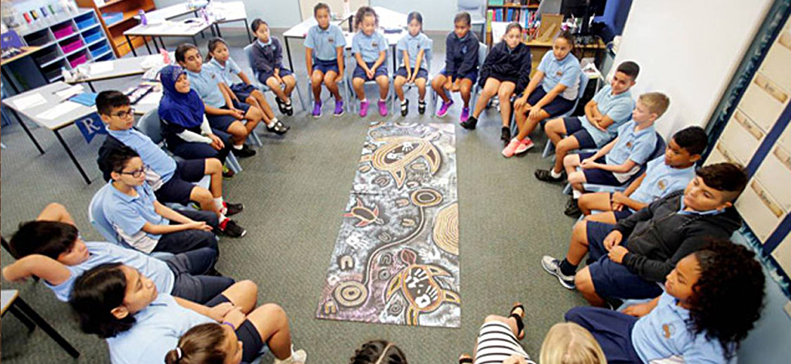 A diverse group of students in blue uniforms sit in a circle, attentively focused on a colorful abstract mural laid out on the floor, fostering a sense of community and learning.