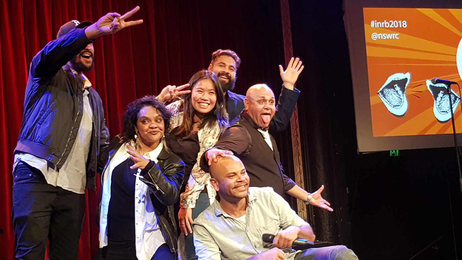 Six people pose humorously on a stage with red curtains. They smile, make peace signs, and gesture lively. A colorful screen displays #inrb2018.