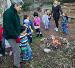 A group of children and two adults gather around a small campfire outdoors. The children appear curious and engaged, dressed warmly in jackets.