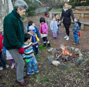 A group of children and two adults gather around a small campfire outdoors. The children appear curious and engaged, dressed warmly in jackets.