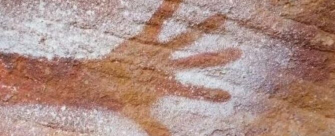 Ancient rock art showing a hand stencil on a reddish-brown stone surface, outlined in white pigment, conveying a connection to past cultures.