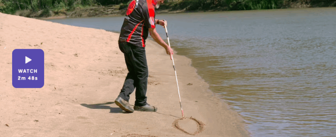 A man wearing a patterned shirt and headband uses a cane to draw symbols in the sand along a riverbank, with trees lining the opposite shore.