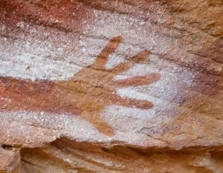 Ancient rock art showing a hand stencil on a reddish-brown stone surface, outlined in white pigment, conveying a connection to past cultures.