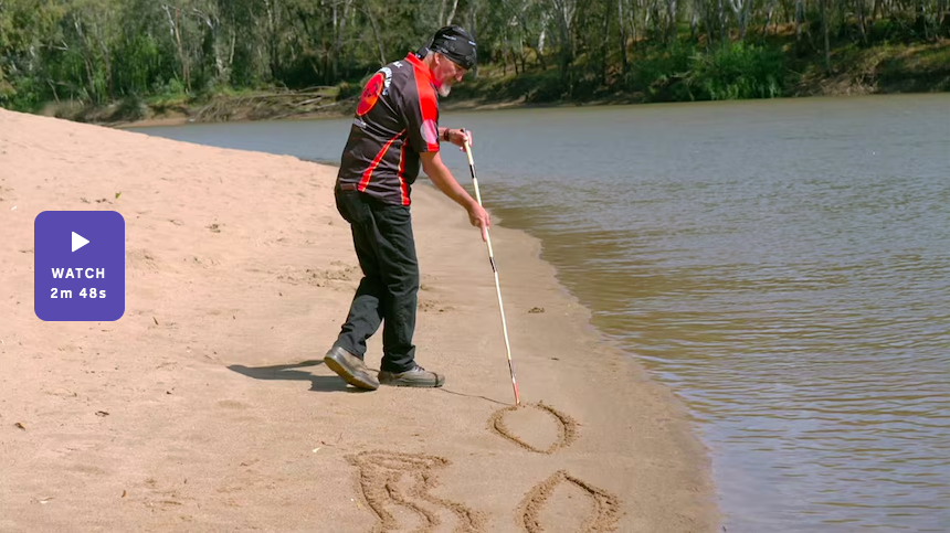 A man wearing a patterned shirt and headband uses a cane to draw symbols in the sand along a riverbank, with trees lining the opposite shore.