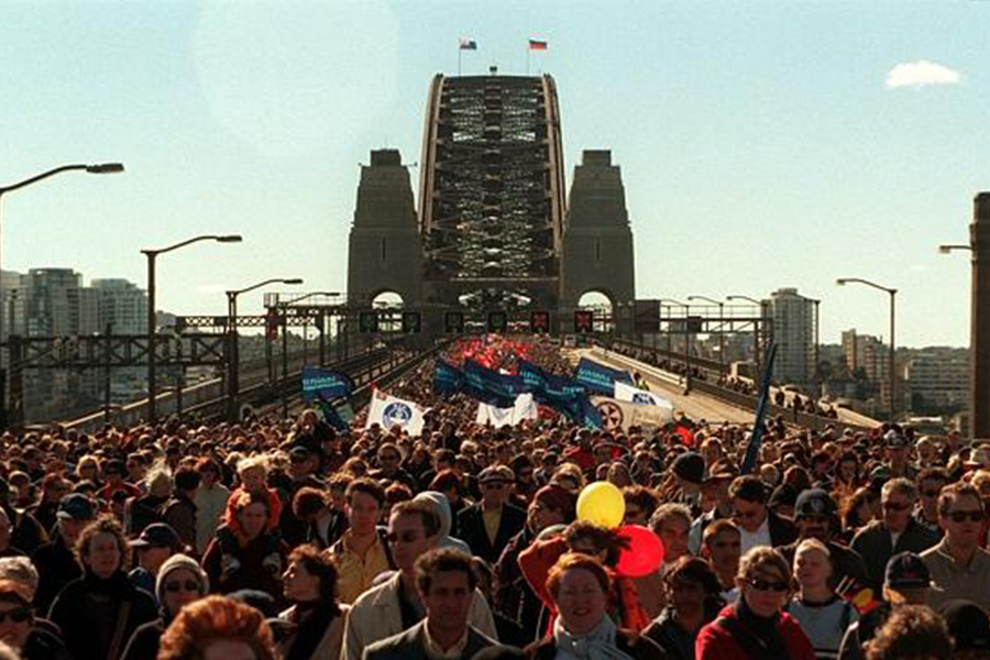 A large crowd marches across the Sydney Harbour Bridge under a clear blue sky. People hold banners and signs. The atmosphere is energetic and hopeful.