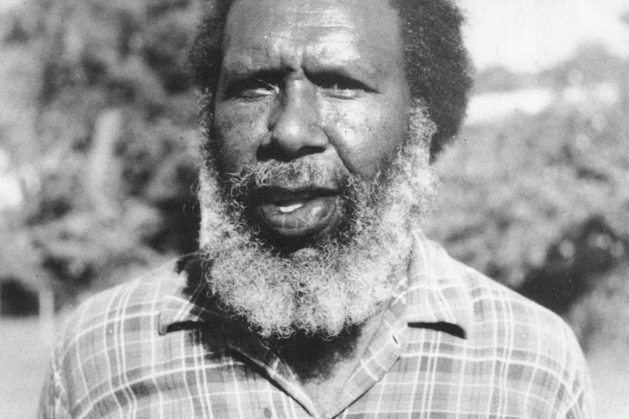 Black and white photo of a man with a thick beard and checkered shirt. He stands outdoors with trees blurred in the background, appearing thoughtful.
