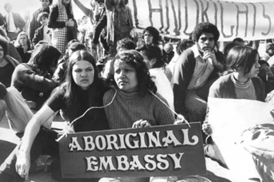 Black and white photo of a protest with people seated, holding a sign reading 'Aboriginal Embassy.' Background banner says 'Land Rights' conveying determination.