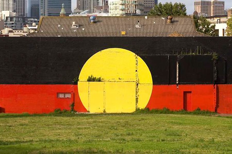 A mural of the Aboriginal flag is painted on a wall, with a black top half, red bottom half, and a large yellow circle in the center. Urban buildings are in the background, with grass in the foreground. The mural conveys a sense of pride and cultural significance.