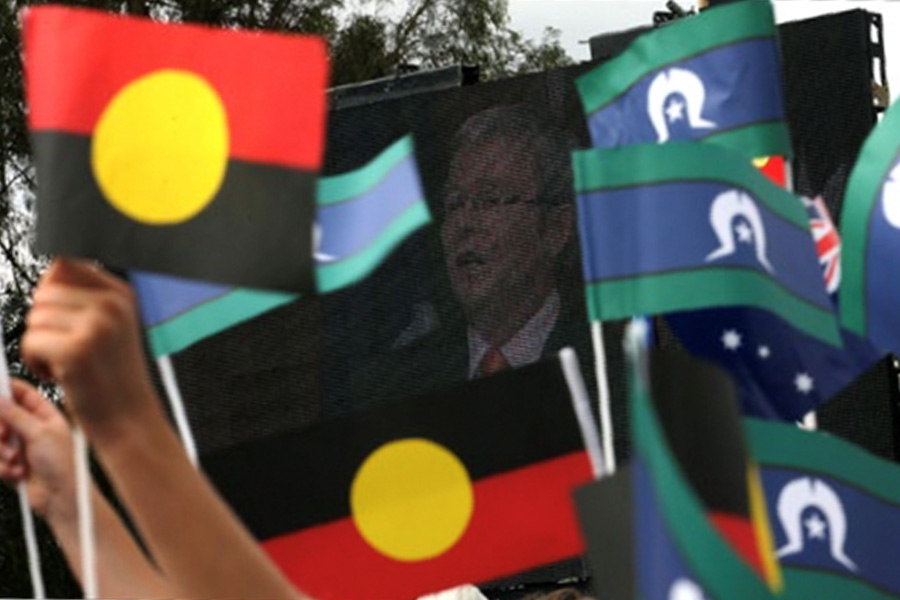 People wave Aboriginal and Torres Strait Islander flags in the foreground, with a large screen in the background showing a person speaking, suggesting a public event.