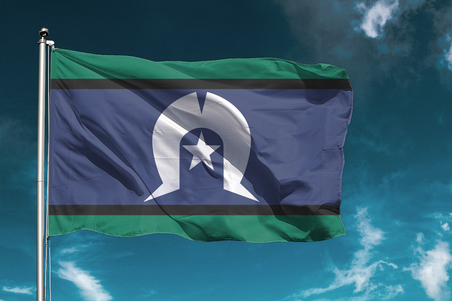 The image shows the Torres Strait Islander flag waving against a blue sky. The flag features green, blue, black, and white colors with a star and headdress symbol.