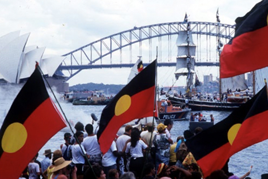 A crowd waves Aboriginal flags near Sydney Harbour, with the Sydney Opera House and Harbour Bridge in the background. A festive, spirited atmosphere.