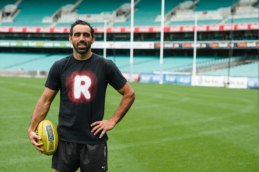 A man stands on a football field holding a yellow ball. He wears a black shirt with a red and white 'R'. The stadium is empty, conveying a calm atmosphere.