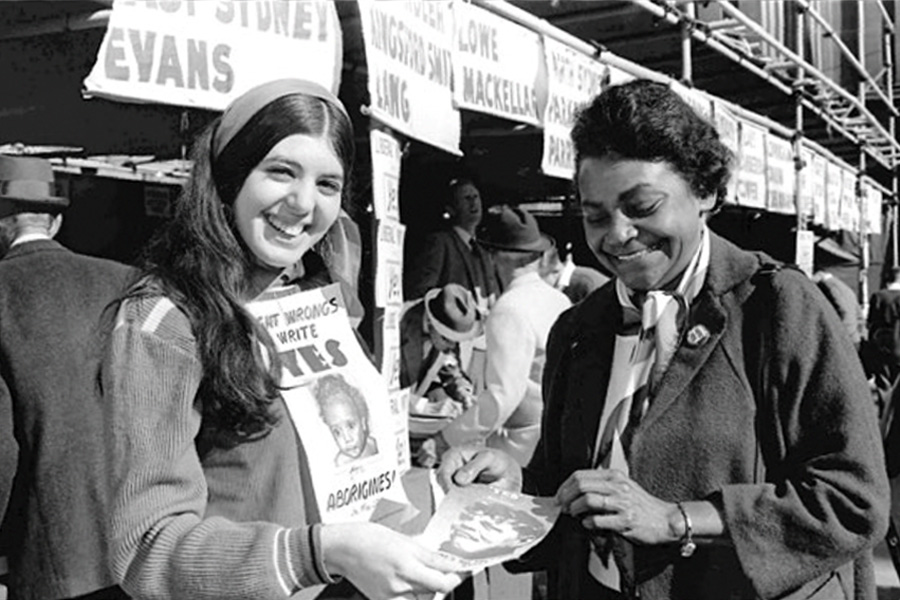 Two women smile at each other, one holding a pamphlet. Behind them are signs and people, indicating a lively atmosphere. The mood is positive and engaged.